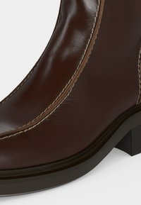 Gros plan sur l'avant et le côté d'une botte en cuir marron foncé avec des coutures visibles et un petit talon bloc.
