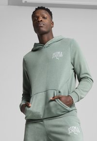 Sudadera verde claro con bolsillo canguro, con el logo "PUMA sports club" en blanco, hecha de un tejido liso y texturizado.