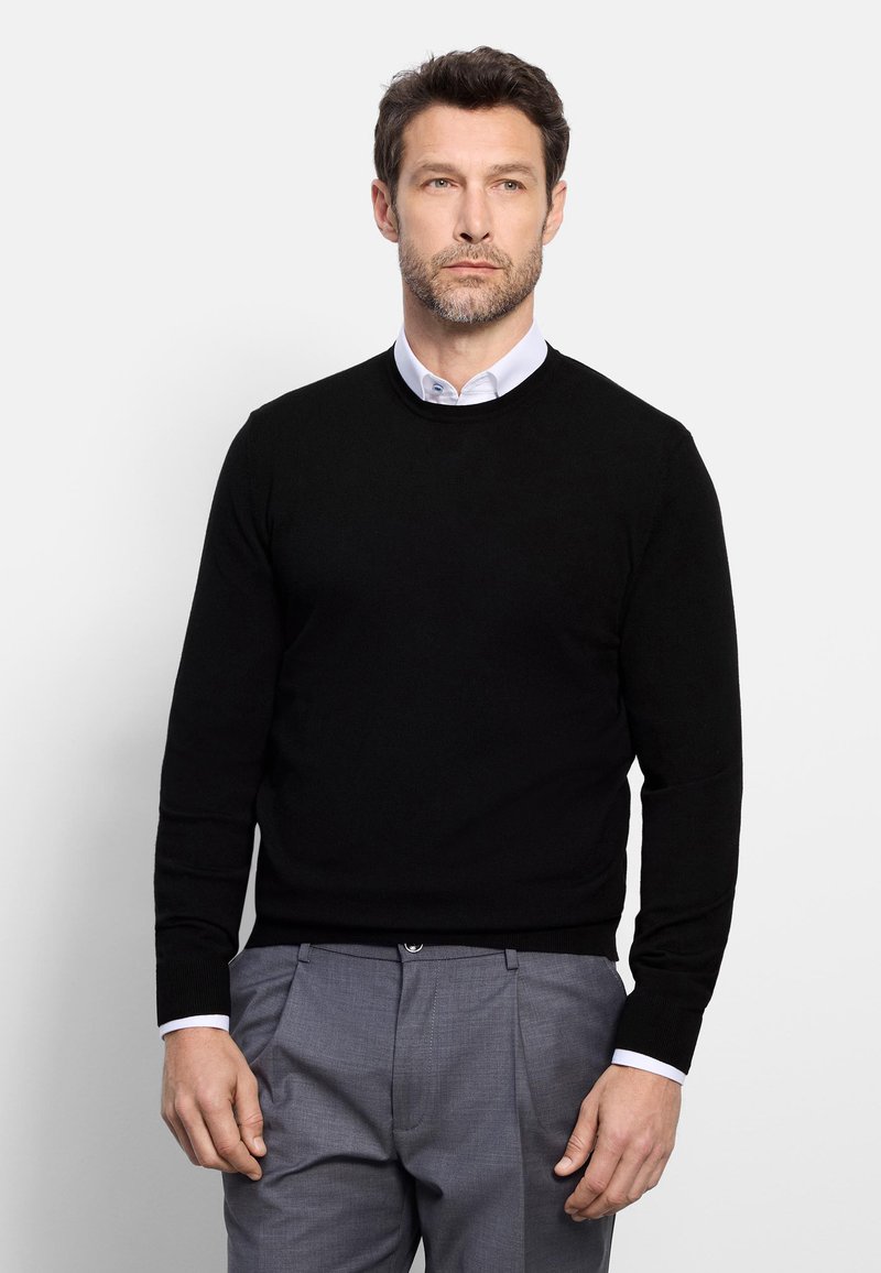 Man with short dark hair and light beard wearing black sweater over white collared shirt and gray pants, standing against plain background.