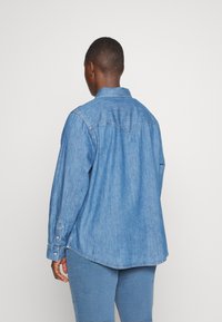 Camicia di jeans blu con maniche lunghe, polsini a bottoni, colletto a punta e dettaglio della yoke posteriore. Il tessuto ha una consistenza morbida e liscia.