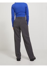 Persona in maglietta lunga blu a maniche, pantaloni larghi grigi scuro con righe, e scarpe nere robuste, in piedi di spalle rispetto all'osservatore su sfondo bianco.