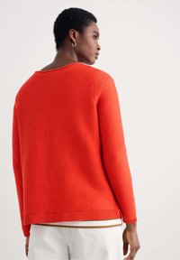 Maglione a coste rosso con scollatura ampia, maniche lunghe e orlo dritto, abbinato a pantaloni bianchi. Texture morbida e vestibilità casual.