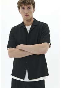 Fekete, rövid ujjú póló texturált mintával, gombos elülső záródással és kontrasztos fehér mandzsettákkal. Fehér kerek nyakú t-shirt-re van húzva.