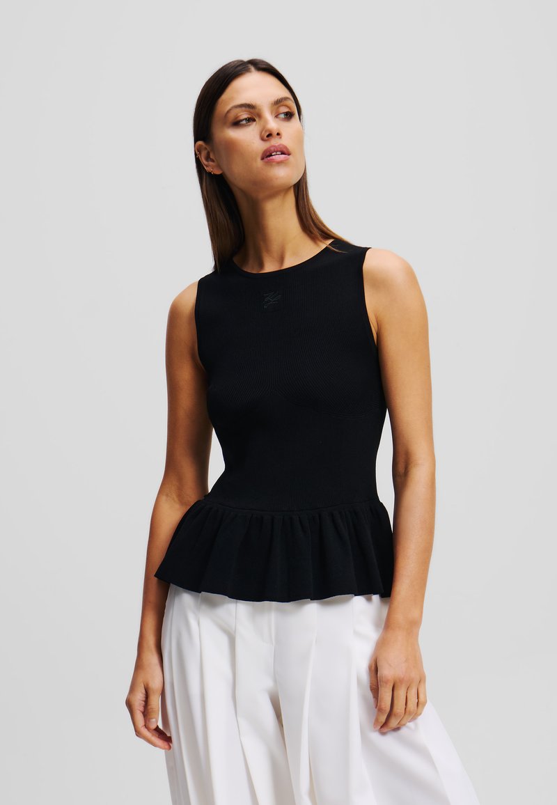 Černý, žebrovaný peplum top s kulatým výstřihem a vrstveným volánkovým lemem, spárovaný s bílými plisovanými kalhotami; decentní branding na hrudi.