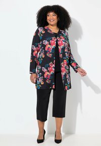 Bloemige cardigan met lange mouwen, zwarte achtergrond, levendige roze en blauwe bloemen, gecombineerd met zwarte cropped broeken en zwarte hakken.