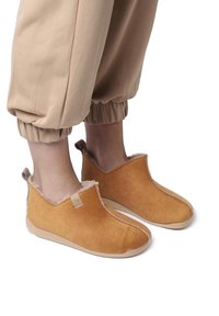 Slip-on slippers in a warm tan color, featuring a soft suede texture, a faux fur lining, and a low-profile design with a rubber sole.