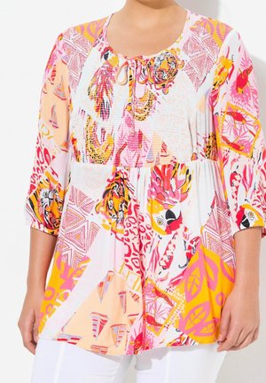Femme portant une tunique colorée à motifs avec des perroquets, des feuilles et des motifs géométriques dans des tons rose, jaune, orange et blanc.