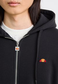 Schwarze Sweatjacke mit Reißverschluss aus Baumwolle. Verfügt über ein rot-orange gesticktes Logo auf der linken Brust und einen metallischen Reißverschluss mit einem dekorativen Zipper.