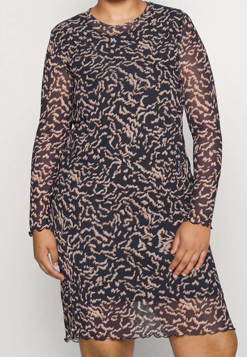 Femme portant une robe noire à manches longues, longueur genou, en tissu transparent avec des motifs abstraits ondulés beiges, ourlet et poignets festonnés.