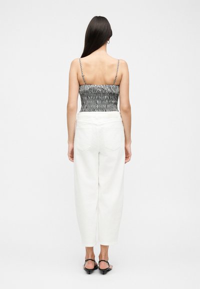 Γυναίκα στέκεται με την πλάτη προς την κάμερα, φοράει ασπρόμαυρο καρό αμάνικο τοπ, άσπρο παντελόνι με ψηλή μέση και μαύρες γόβες.