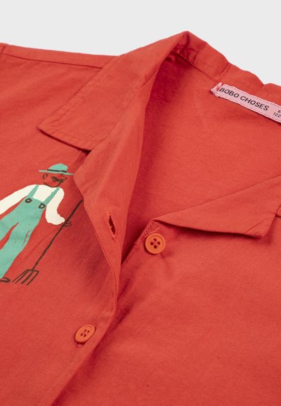 Chemise boutonnée couleur rouille avec col, arborant une petite illustration d'un fermier tenant une fourche, portant une salopette verte et un chapeau.