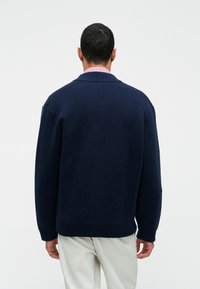 Tumedat sinise värvi kudumisjärgmise särgi, millel on soonikuga krae ja äär, laia lõikega ja langevate õlgadega, nähtuna selja tagant.