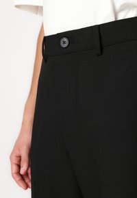 Svarta skräddarsydda byxor med en slät yta, med knappstängning, bälteshällor och en sidoficka, delvis synliga tillsammans med en hand.