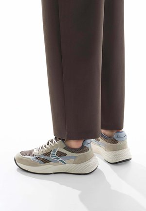 Sneakers hebben een beige en grijze mesh bovenwerk met blauwe accenten, een gewatteerde witte zool en een gestructureerde zwarte buitenzool.