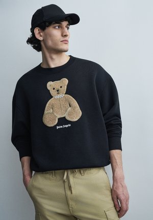 Νέος άνδρας με μαύρο καπέλο, μαύρο φούτερ με σχέδιο αρκουδάκι σε μπεζ χρώμα και μπεζ παντελόνι, κοιτάζοντας προς τα πλάγια μπροστά σε απλό γκρι φόντο.