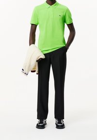 Osoba v jasně zeleném polo tričku, černých kalhotách, černých teniskách, držící bílý svetr s barevnými pruhy v jedné ruce.