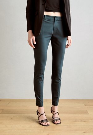 Pantaloni tailored color teal con una texture liscia, taglio slim e aperture alla caviglia, abbinati a tacchi neri con cinturini su un pavimento in legno.