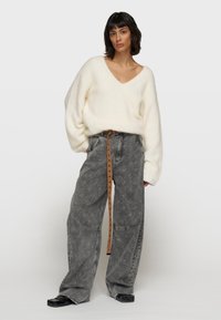 Weißer, flauschiger, übergroßer Pullover mit tiefem V-Ausschnitt, kombiniert mit gemusterten grauen Jeans und einem braunen Gürtel. Schwarze Schuhe runden den Look ab.
