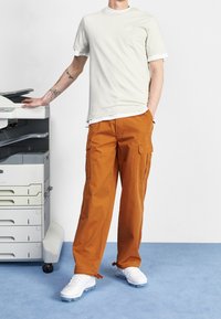 Ljust grå kortärmad skjorta, orange cargobyxor med fickor och vita sportskor, står bredvid en kopiator på blå matta.