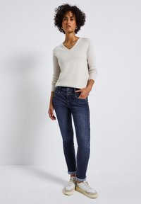 Pull à col en V beige clair avec manches longues, associé à un jean slim bleu foncé et des baskets beiges. Texture lisse, coupe décontractée.