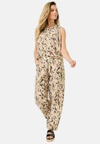 Beige jumpsuit med svarta och rostfärgade lövmönster, elastisk midja, ärmlös design och sidofickor, kombinerad med svarta kilklackar.