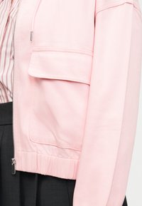 Φωτεινό ροζ μπουφάν με μπροστινό φερμουάρ και μεγάλο τσεπάκι με καπάκι, φορεμένο πάνω από ροζ και άσπρο ριγέ πουκάμισο και σκούρο παντελόνι.