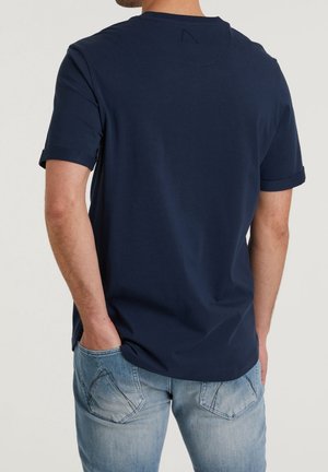 Mężczyzna ubrany w granatowy T-shirt z krótkim rękawem i jasnoniebieskie jeansy, ręce w tylnych kieszeniach, odwrócony tyłem do kamery.