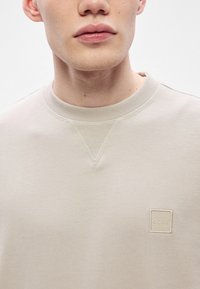 Ανοιχτό μπεζ φούτερ με στρογγυλή λαιμόκοψη, εμφανείς ραφές και μικρό τετράγωνο λογότυπο με την ένδειξη "BOSS" στο στήθος. Μαλακή υφή υφάσματος.