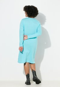 Ein langärmeliges, tealfarbenes Kleid aus weichem Stoff mit lockerem Sitz. Kombiniert mit schwarzen, klobigen Stiefeln mit Absatz.