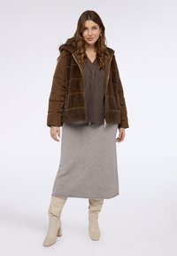 Giacca imbottita marrone con cappuccio, abbinata a un top lavorato a maglia grigio scuro e una lunga gonna grigia. L'outfit è completato da stivali beige al ginocchio.