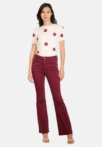 Donna che indossa un maglione a maniche corte color crema con stampe di fiori rossi e pantaloni a campana bordeaux a vita alta, in piedi dritta con un'espressione neutra.