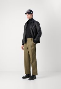 Veste en cuir noir, col roulé noir, pantalon plissé vert olive et chaussures noires. Casquette noire avec le logo "HUGO" en rouge et blanc.