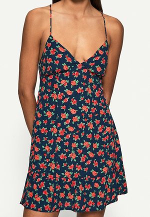 Femme portant une robe sans manches bleu marine avec un motif floral rouge et vert et de fines bretelles spaghetti.