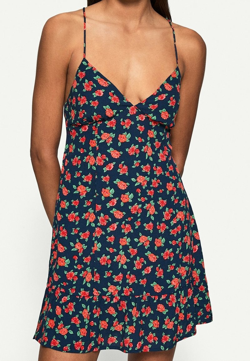 Femme portant une robe sans manches bleu marine avec un motif floral rouge et vert et de fines bretelles spaghetti.