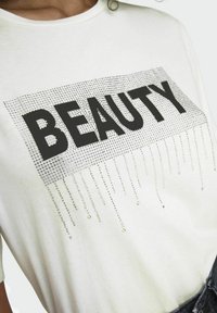 Witte t-shirt met korte mouwen met het woord "SCHOONHEID" in vette zwarte letters. Versierd met zilveren rhinestones in een verlopen patroon onder de tekst.