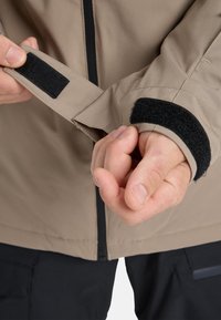 Beige jacket featuring black Velcro cuffs. Close-up of hands adjusting the sleeve, highlighting the smooth fabric and fitted design.