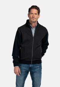 Heren zip-up jas met een donkere textuur op het lichaam en marineblauwe mouwen. Kenmerkt zich door een hoge kraag en zijzakken, gedragen over een patroonshirt.