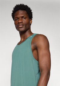 Sleeveless teal tank top, made of textured fabric, featuring a rounded neckline and a small logo detail on the shoulder.