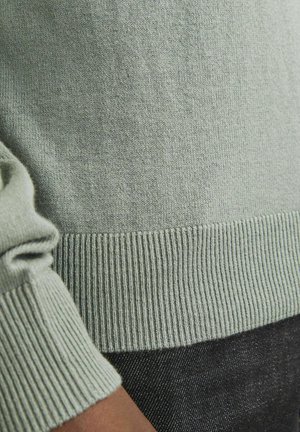 Világos zöld kötött pulóver, bordázott mandzsettákkal és aljjal, puha tapintással. A kép a szövet részleteire és a ujjdesignra összpontosít.