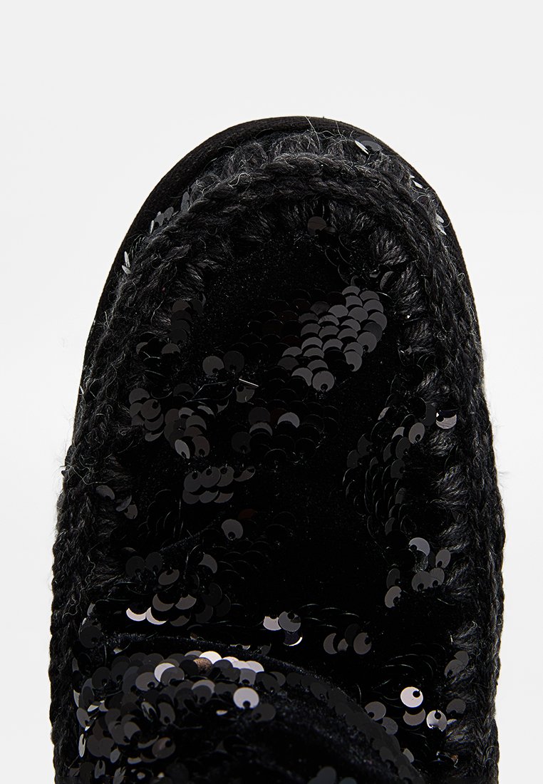 Pantofola nera con tessuto di paillettes, punta arrotondata e bordo intrecciato. La parte superiore è liscia con una texture lucida data dalle paillettes.