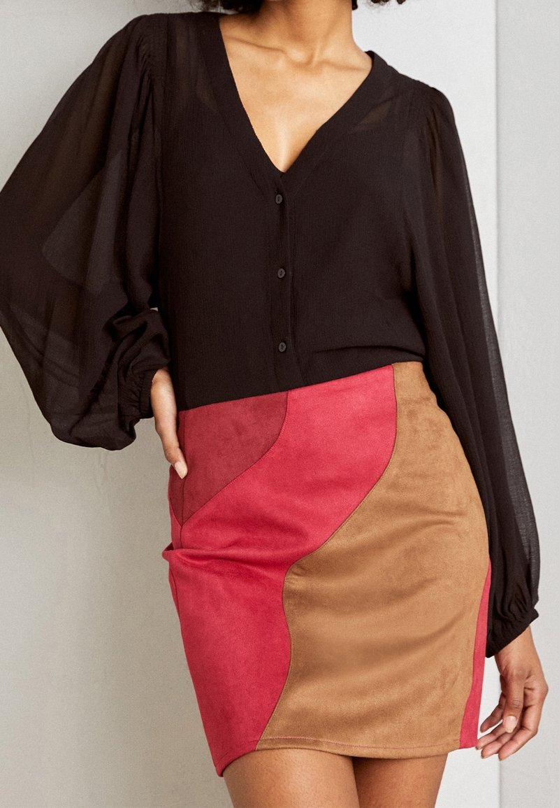 Blouse noire transparente avec boutons sur le devant, associée à une jupe ajustée ornée de panneaux en daim rouge et beige aux motifs courbes.
