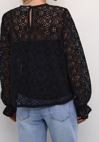 Blouse en dentelle noire à motif floral, avec manches longues et poignets à volants, et une ouverture en forme de goutte d'eau au dos. Portée sur un jean en denim.