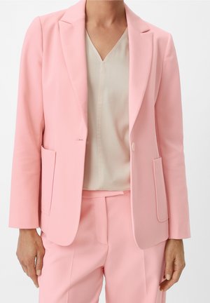 Femme portant un blazer rose pâle cintré et un pantalon assorti sur un chemisier crème à col en V, visible du cou aux hanches.
