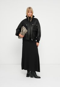 Crna kožna bomber jakna s dva prednja džepa, preko duge crne suknje. Model drži clutch torbicu s uzorkom zmije i nosi crne čizme na petu.