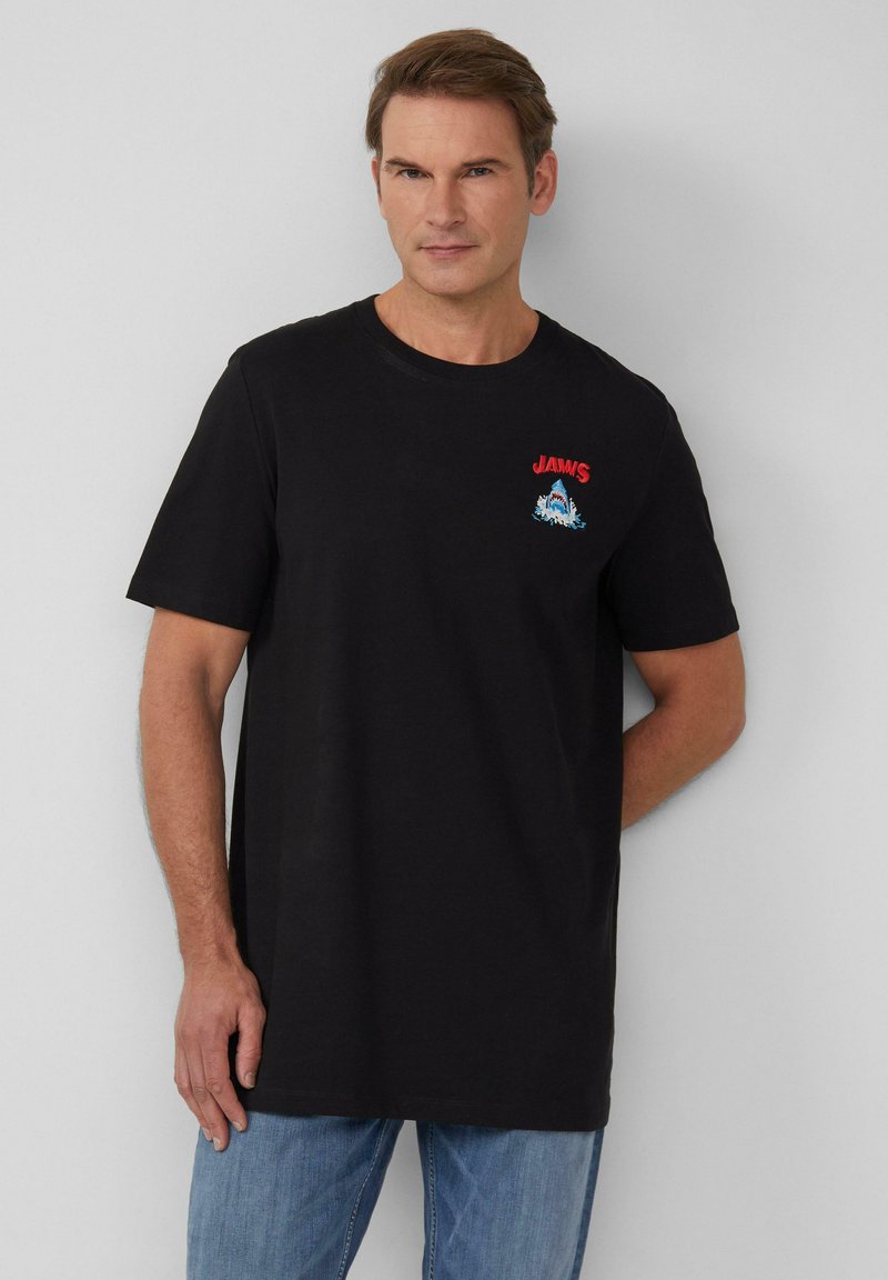 Man in een zwart T-shirt met een rood "JAWS" logo en haaienafbeelding op de borst, gecombineerd met blauwe jeans, staand tegen een effen achtergrond.