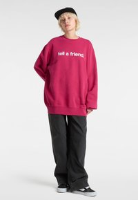 Fuchsie v oversize mikině s bílým nápisem "řekni to příteli", v kombinaci s černými kalhotami s širokými nohavicemi a černými teniskami. Kšiltovka na hlavě.