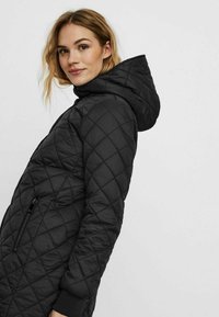 Cappotto imbottito trapuntato nero con cappuccio, caratterizzato da motivi a diamante, tasche laterali e polsini a costine. Il materiale sembra liscio e leggero.