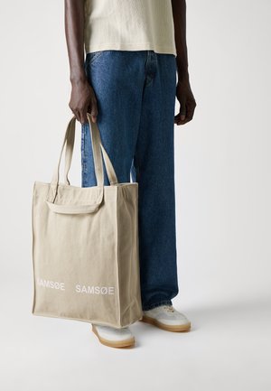 Canvas tote bag in beige with white "SAM SøE" logo, featuring soft texture, rectangular shape, and sturdy handles, held by a person.