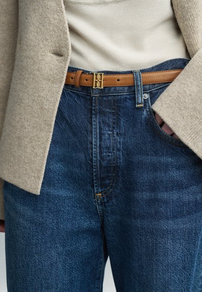 Τζιν σε σκούρο μπλε με διακριτή υφή, συνδυασμένα με ένα μπεζ μάλλινο σακάκι. Η καφέ δερμάτινη ζώνη διαθέτει αγκράφα σε χρυσό τόνο.