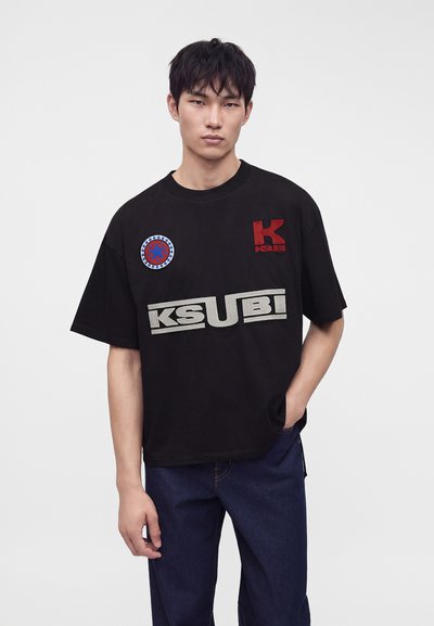 Νέος άνδρας που φοράει μαύρο t-shirt Ksubi με κόκκινα και λευκά λογότυπα και σκούρο μπλε τζιν, στέκεται με το ένα χέρι στην τσέπη σε λευκό φόντο.